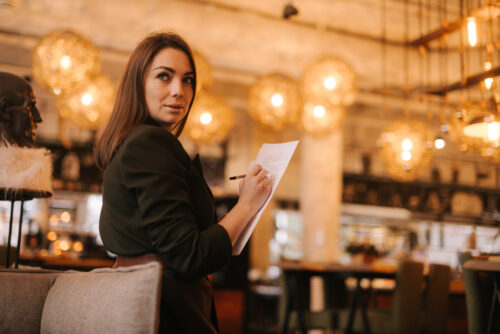 Business young woman wearing stylish formal clothes in modern luxury restaurant holding paper documents and pen in her hands. Business lady lady in suit posing in cafe.